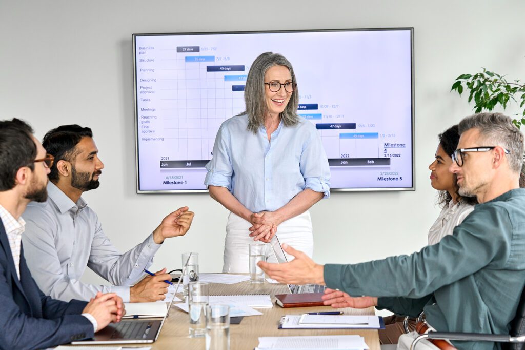 Female standing in front of a white board with 4 seated individuals