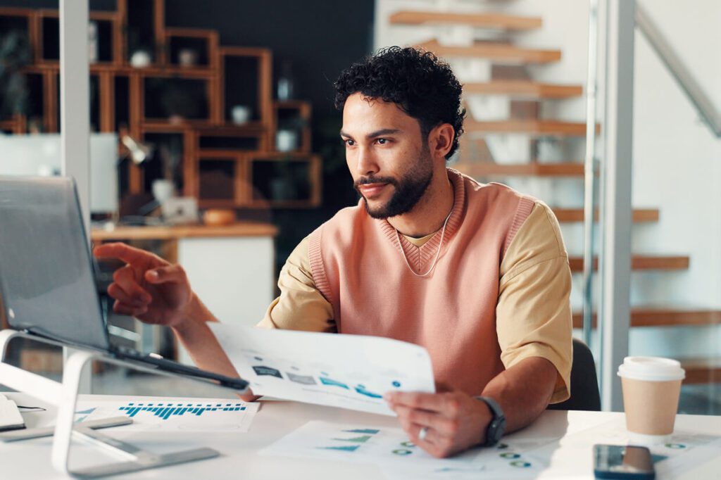 Man seated around a desk looking and pointing to a laptop screen holding a sheet in one hand