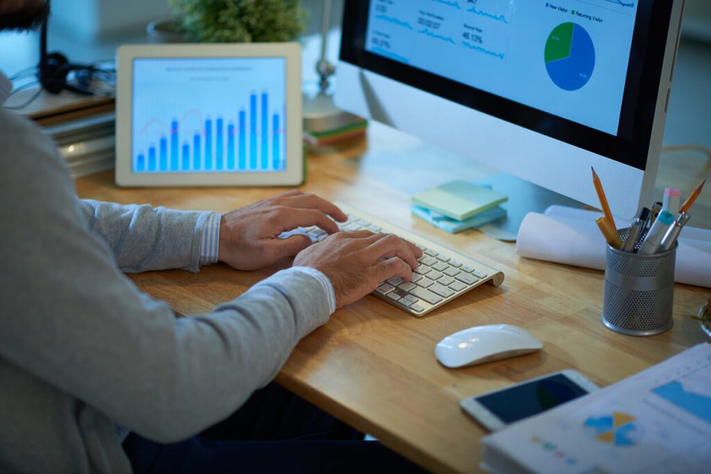 Male typing on a keyboard with a tablet, phone and desktop monitor around a desk