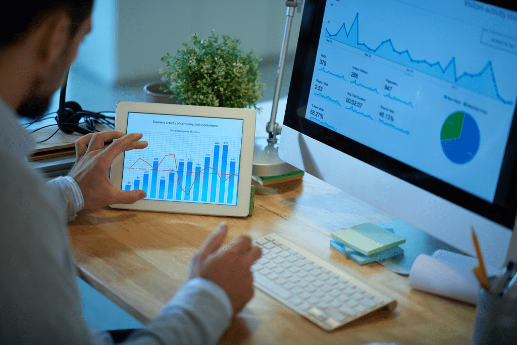 Male touching a tablet screen with a bar and line graph seated in from of a computer monitor with charts and numbers